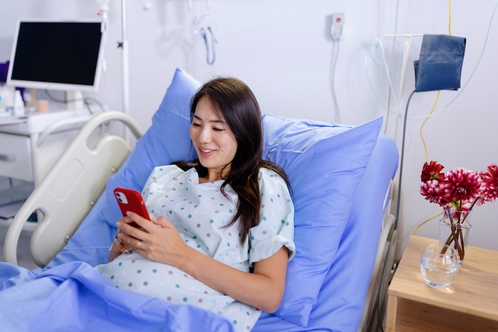 Woman talking to her friends and family on her phone while smiling in a hospital bed at a fetal center next to a bouquet of flowers.