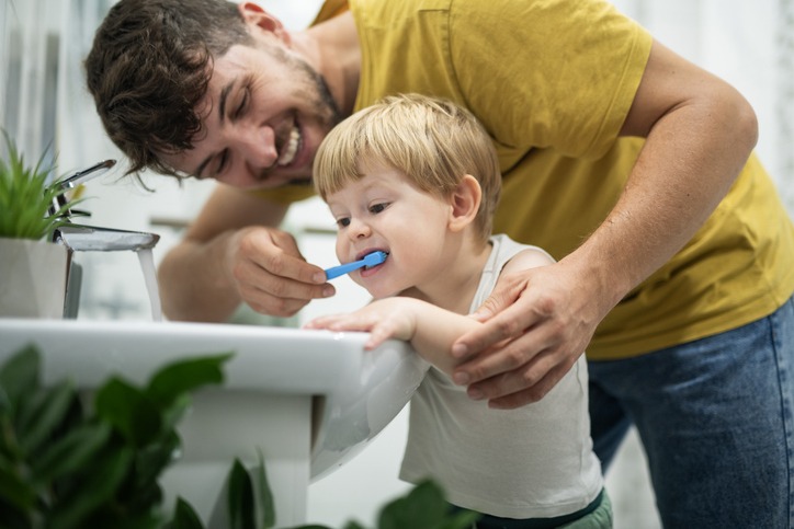 Father teaching his toddler to wash his teeth in the bathroom.