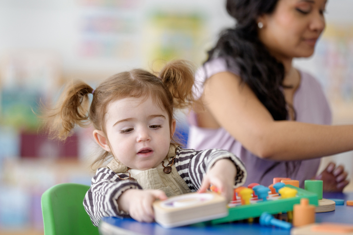 A preschool-aged girl explores a colorful learning toy at a small table, with a smiling caregiver nearby. The bright, friendly classroom setting highlights early learning, play, and focus.