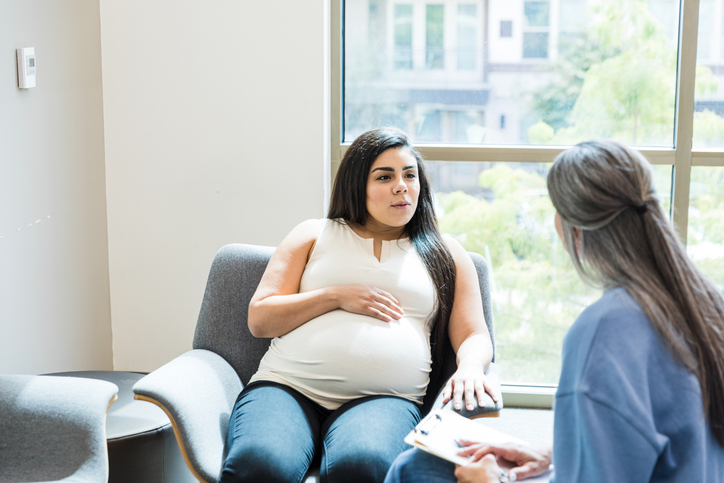 A pregnant woman sitting down in a chair talking with her doctor.
