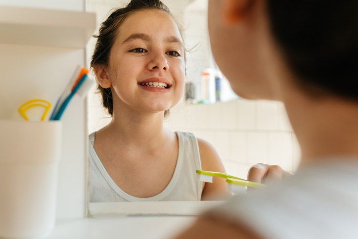 A young girl in a bathroom, looking herself in the mirror while holding a toothbrush after brushing her teeth with fluoride toothpaste.